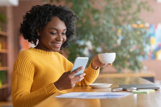Mujer sonriendo y mirando su teléfono inteligente mientras bebe café