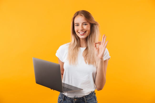 Mujer joven sonriente que muestra un buen gesto con la mano mientras sostiene una computadora portátil.